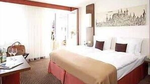 Una habitación de hotel con cama, escritorio, silla, bolso y copa de vino.
