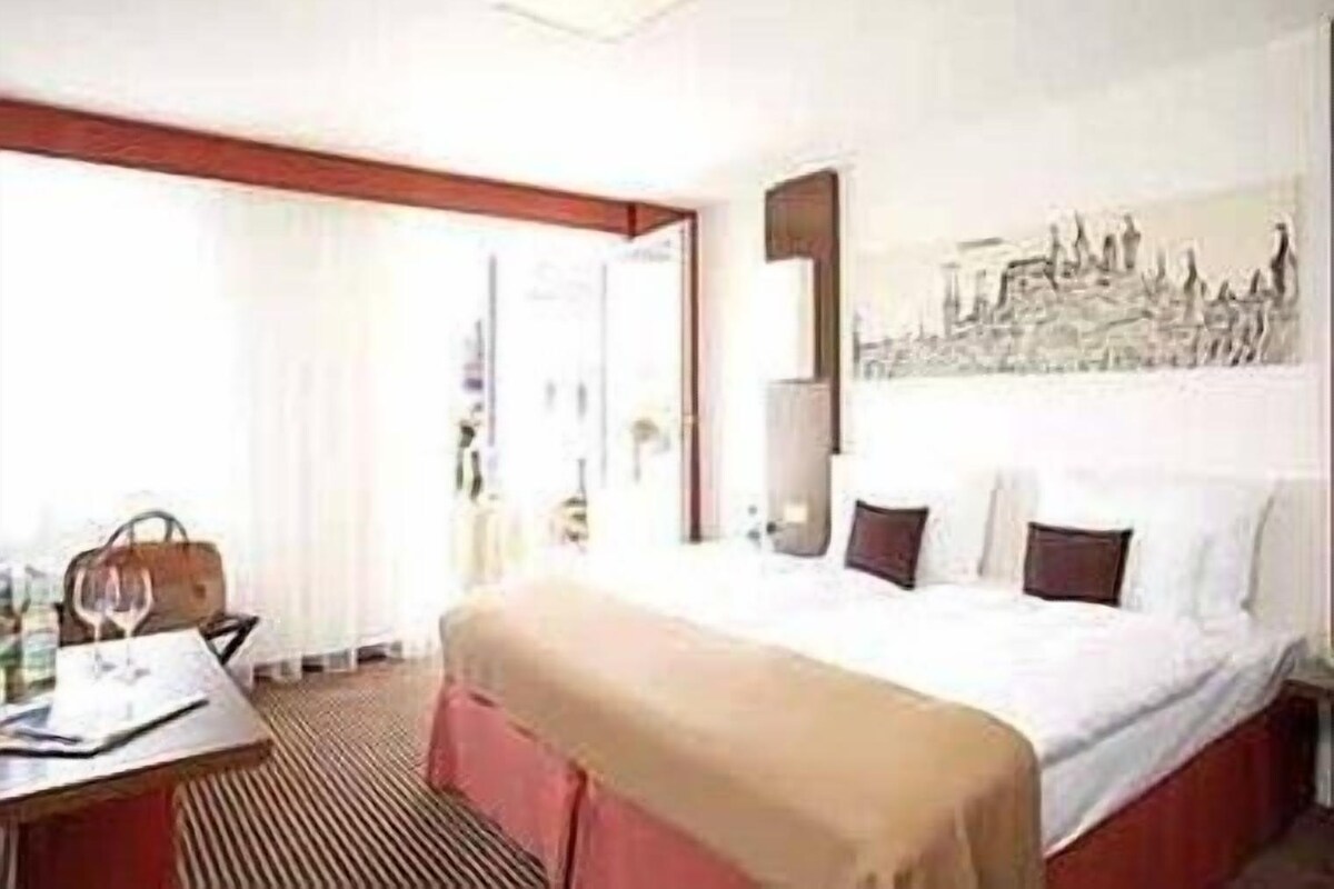 Una habitación de hotel con cama, escritorio, silla, bolso y copa de vino.