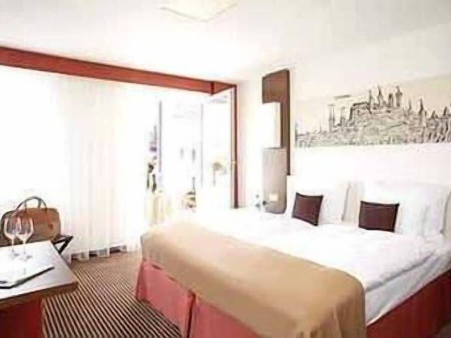 A hotel room with a bed, a desk with a chair, a handbag, and a wine glass.