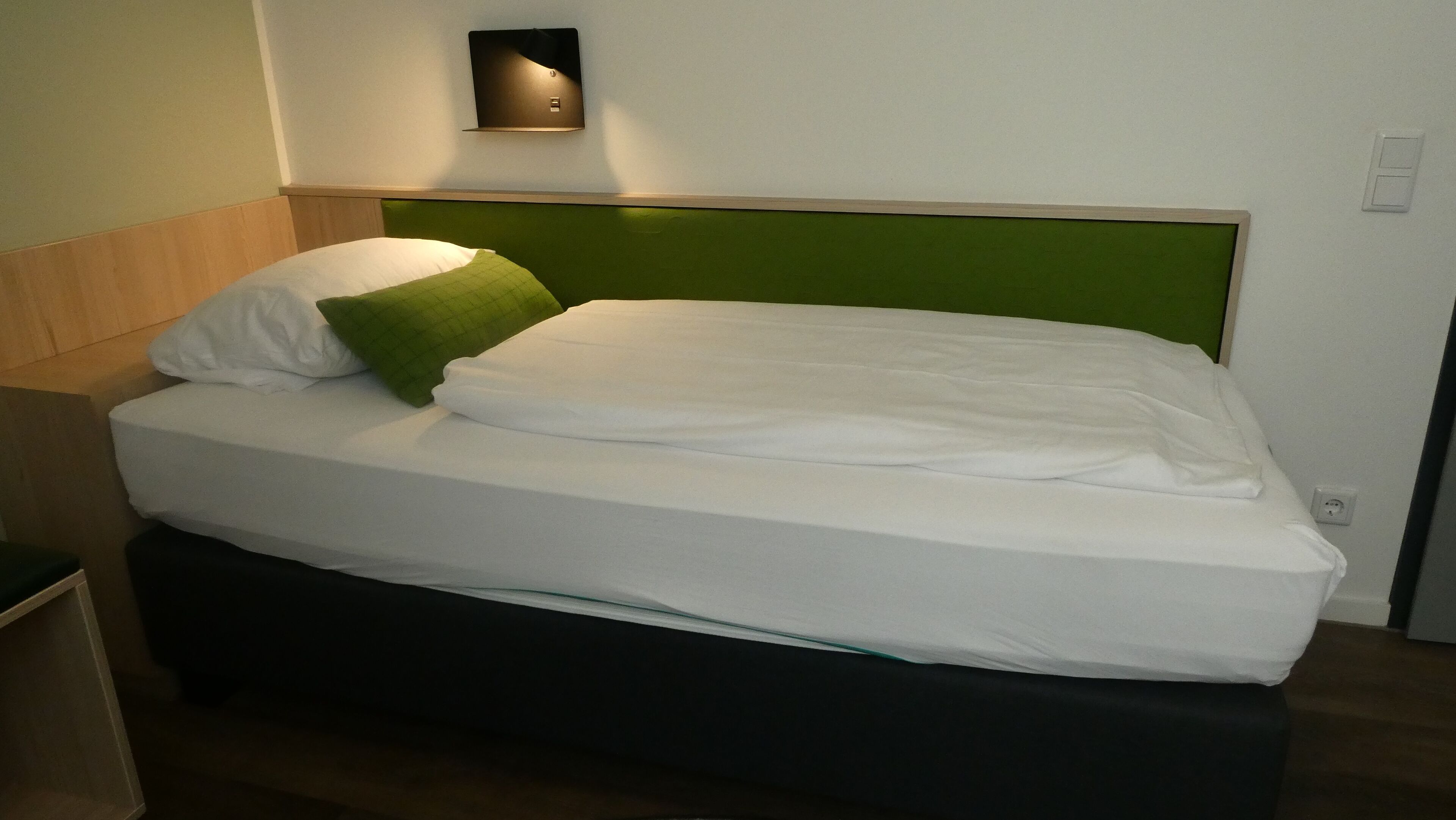 A bed with white bedding and a green headboard, a wall-mounted light fixture, and a power outlet on the wall.
