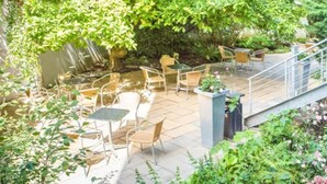 An outdoor patio area with multiple tables and chairs, surrounded by lush greenery and a small water feature. - CVJM Düsseldorf Hotel & Tagung (Düsseldorf)