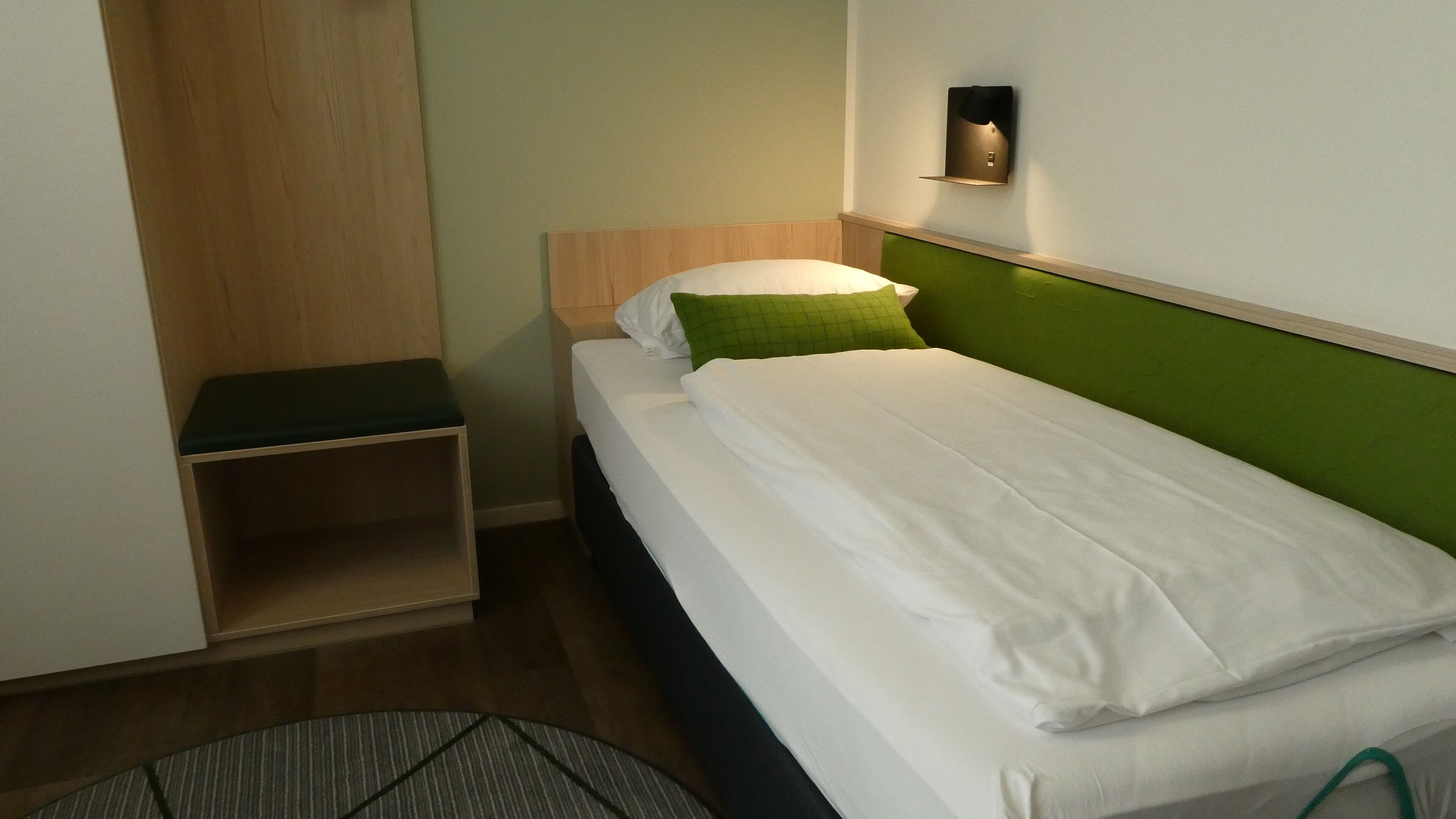 A single bed with white bedding and a green pillow, a bedside table, and a wall-mounted light fixture.