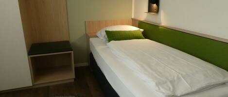 A single bed with white bedding and a green pillow, a bedside table, and a wall-mounted light fixture.