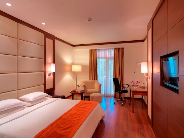 A hotel room with a large bed, a desk with a chair, a TV mounted on the wall, and a window with curtains.