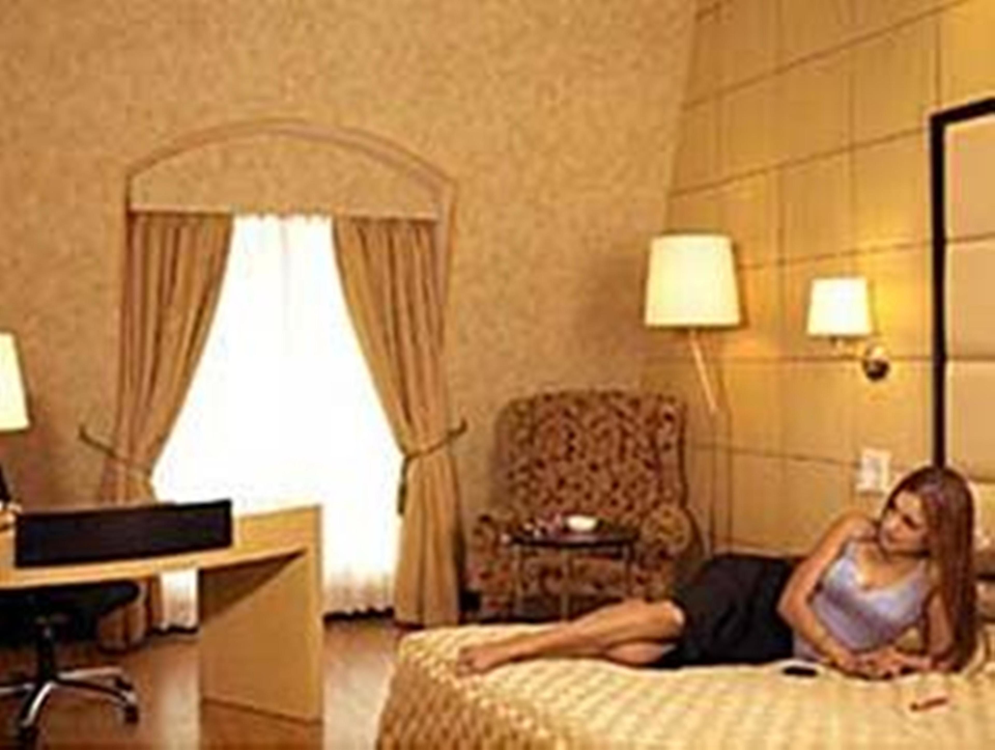 A hotel room with a bed, a desk, a chair, a lamp, and a window with curtains.