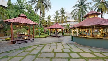 Relax in a serene garden setting surrounded by lush greenery and palm trees, with shaded seating areas under wooden gazebos.
