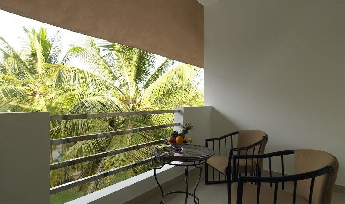 A balcony with a view of palm trees, a round table with a fruit bowl, and two chairs.