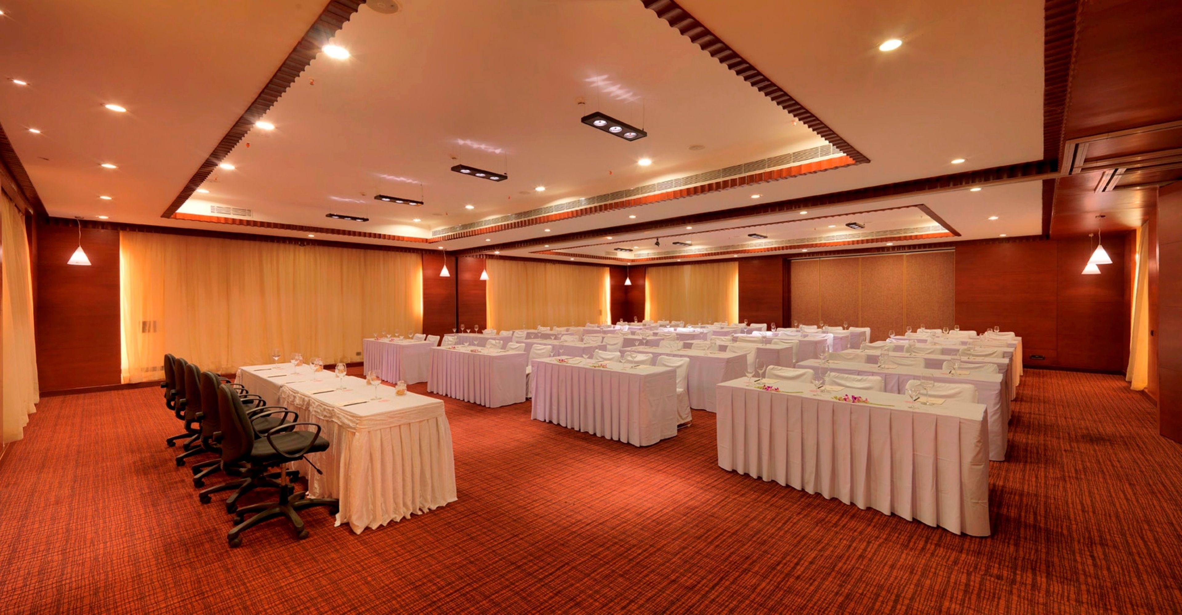 Un salón de conferencias con varias mesas redondas, cada una con mantel blanco y sillas, y piso cubierto de alfombra.