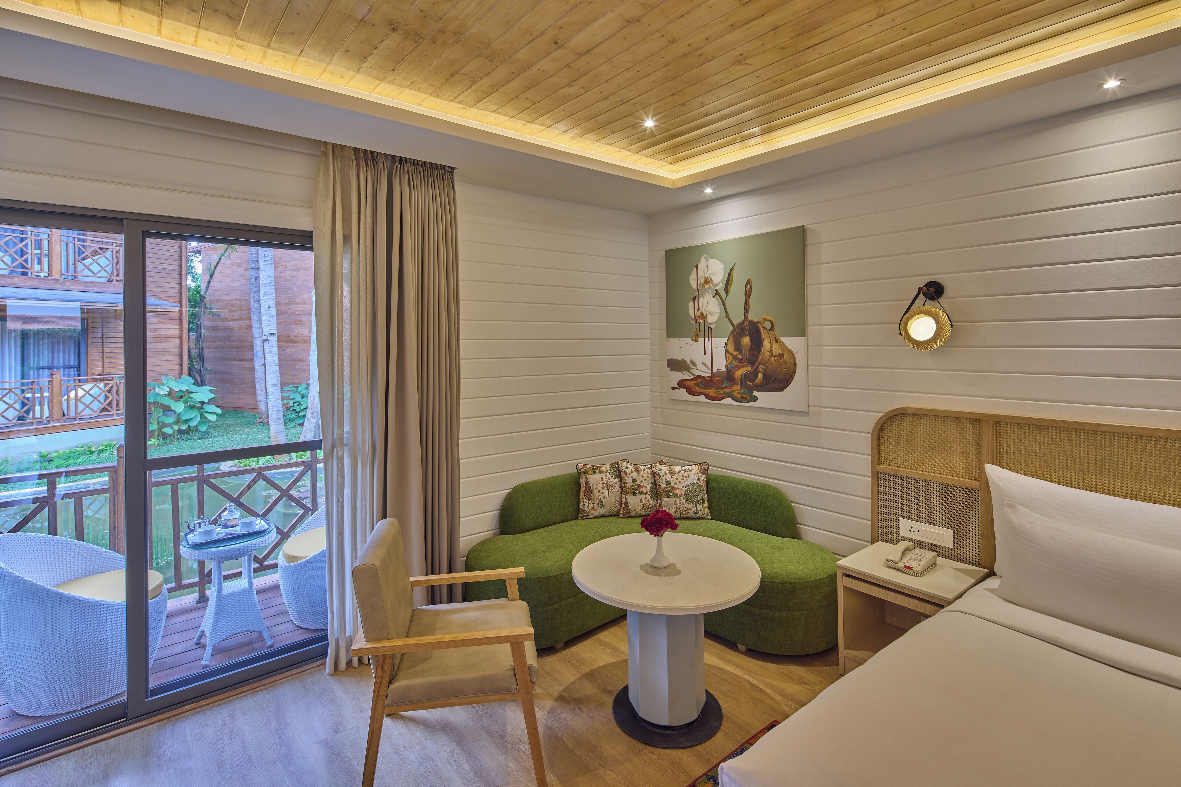 A hotel room with a large sliding glass door leading to an outdoor area, a green sofa, a round table with a vase of flowers, a bed with white bedding, and a wall-mounted lamp.