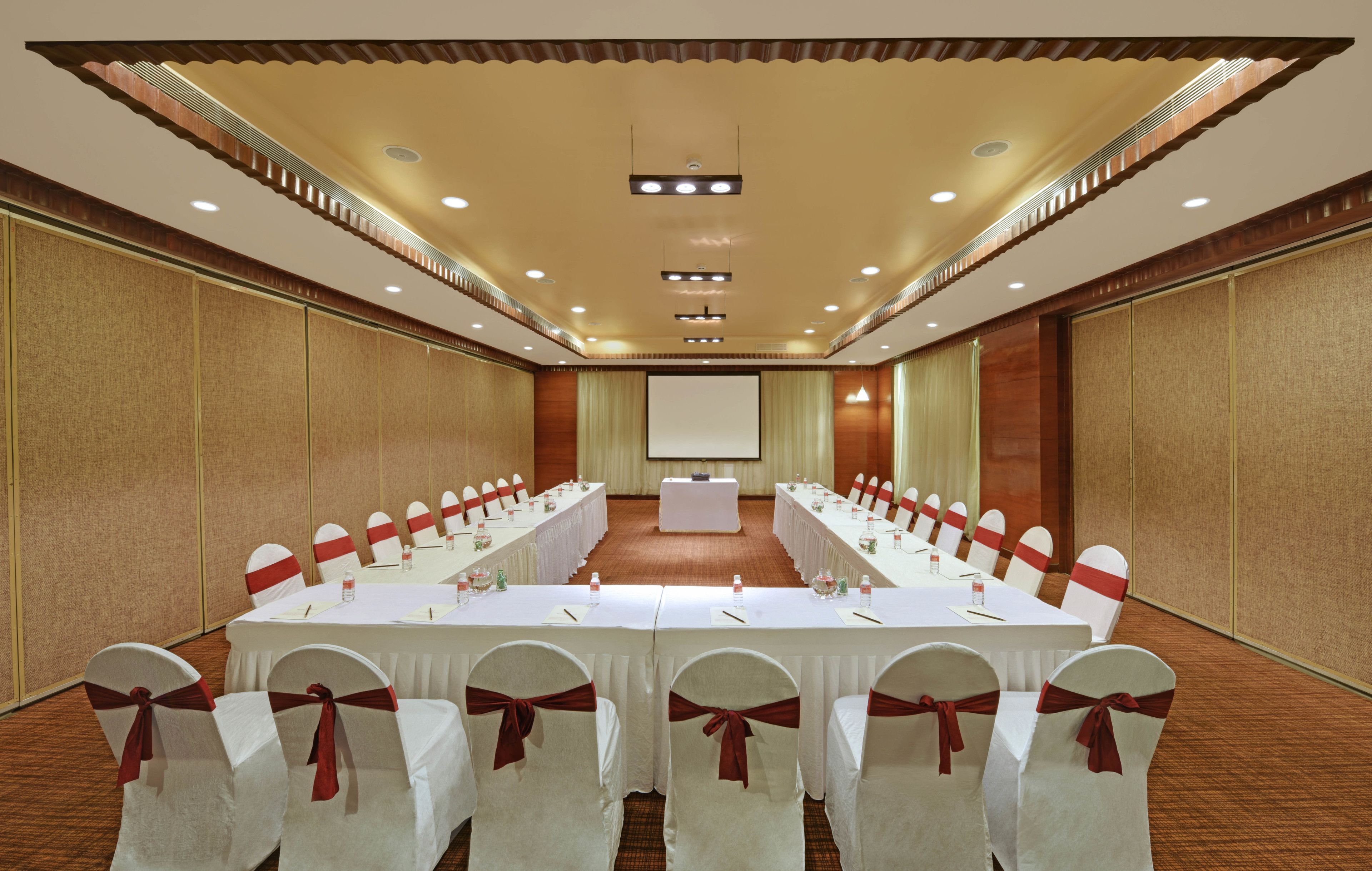 A conference room with long tables, white chairs with red sashes, and a large screen at the front.