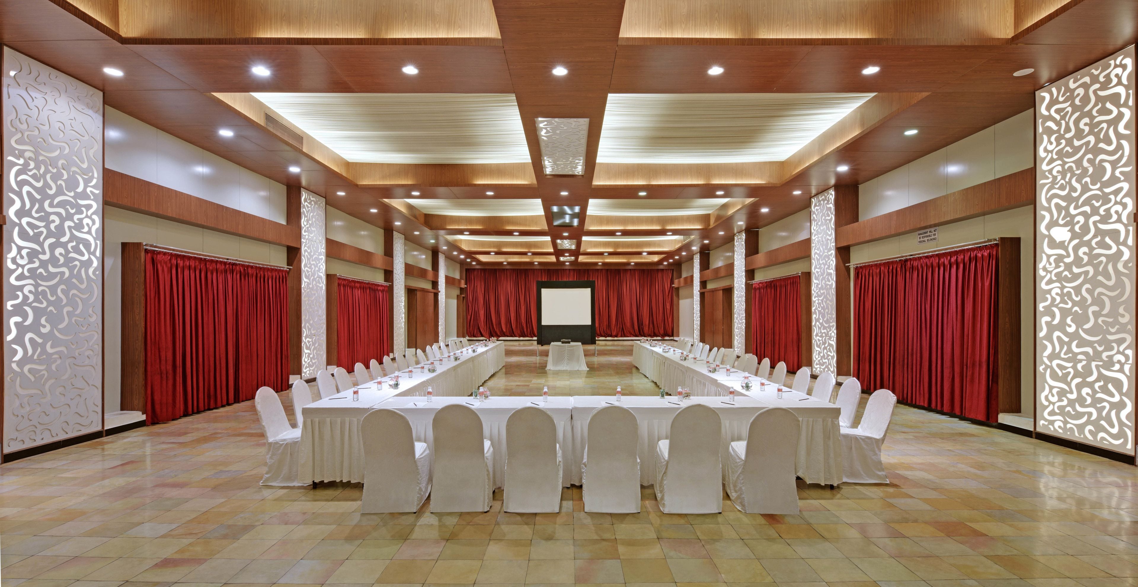 A conference room with long tables, white chairs, and red curtains.