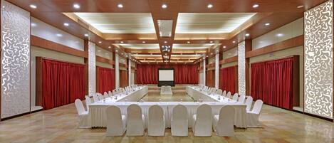 A conference room with long tables, white chairs, and red curtains.