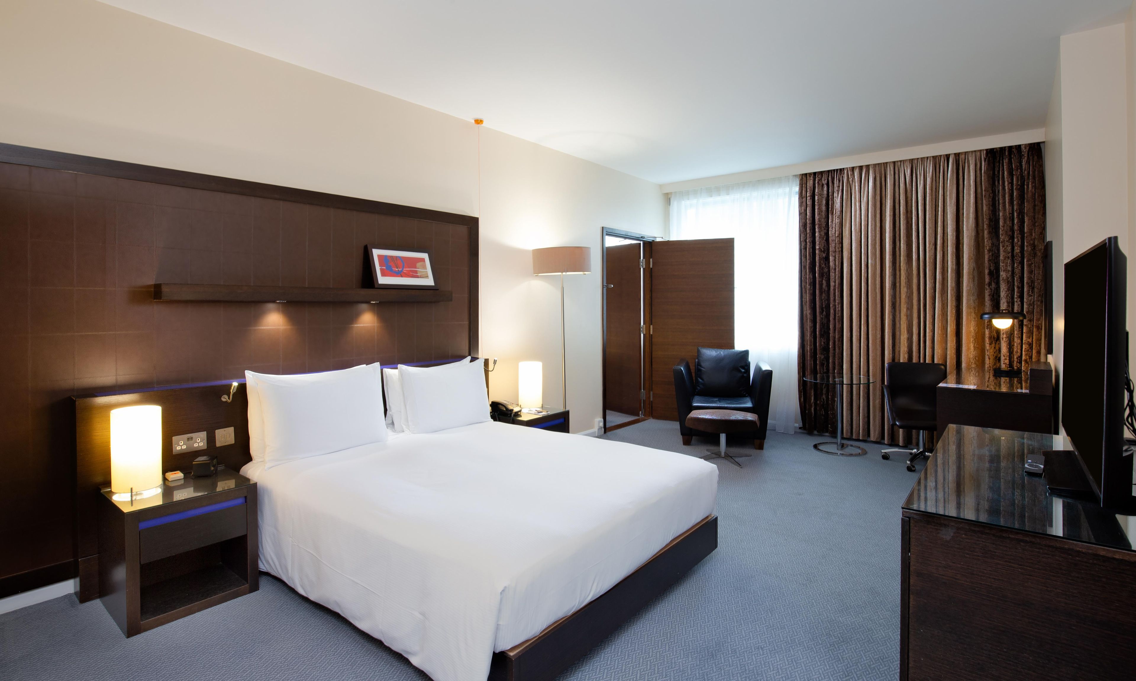 A hotel room with a large bed, bedside tables, a desk with a chair, a TV, and a window with curtains.