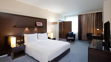 A hotel room with a large bed, bedside tables, a desk with a chair, a TV, and a window with curtains.