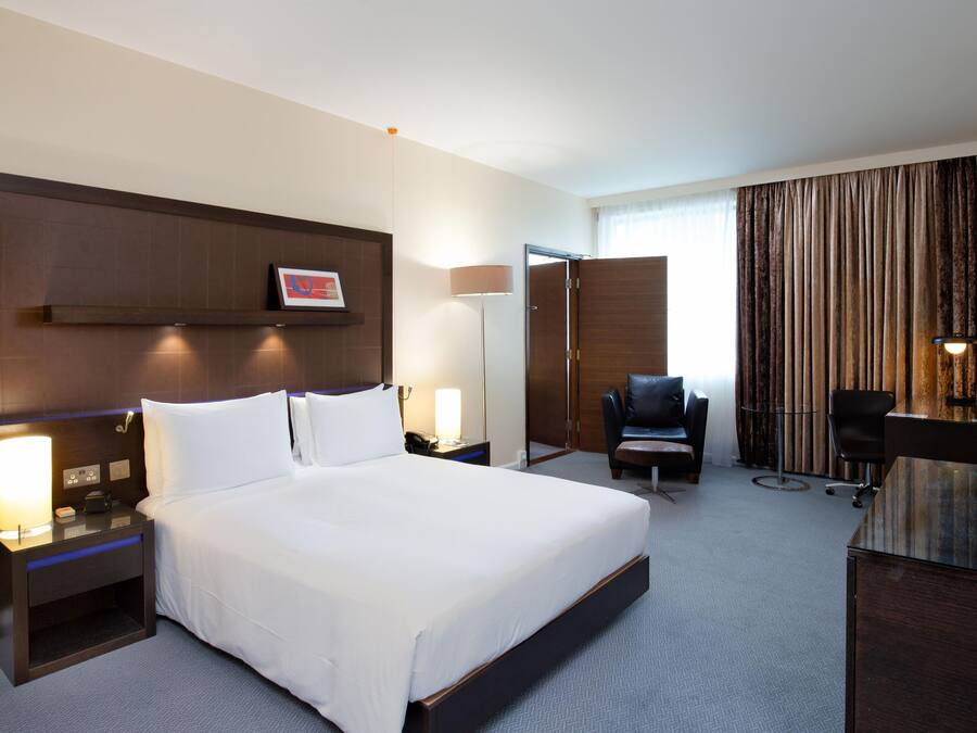 A hotel room with a large bed, bedside tables, a desk with a chair, a TV, and a window with curtains.