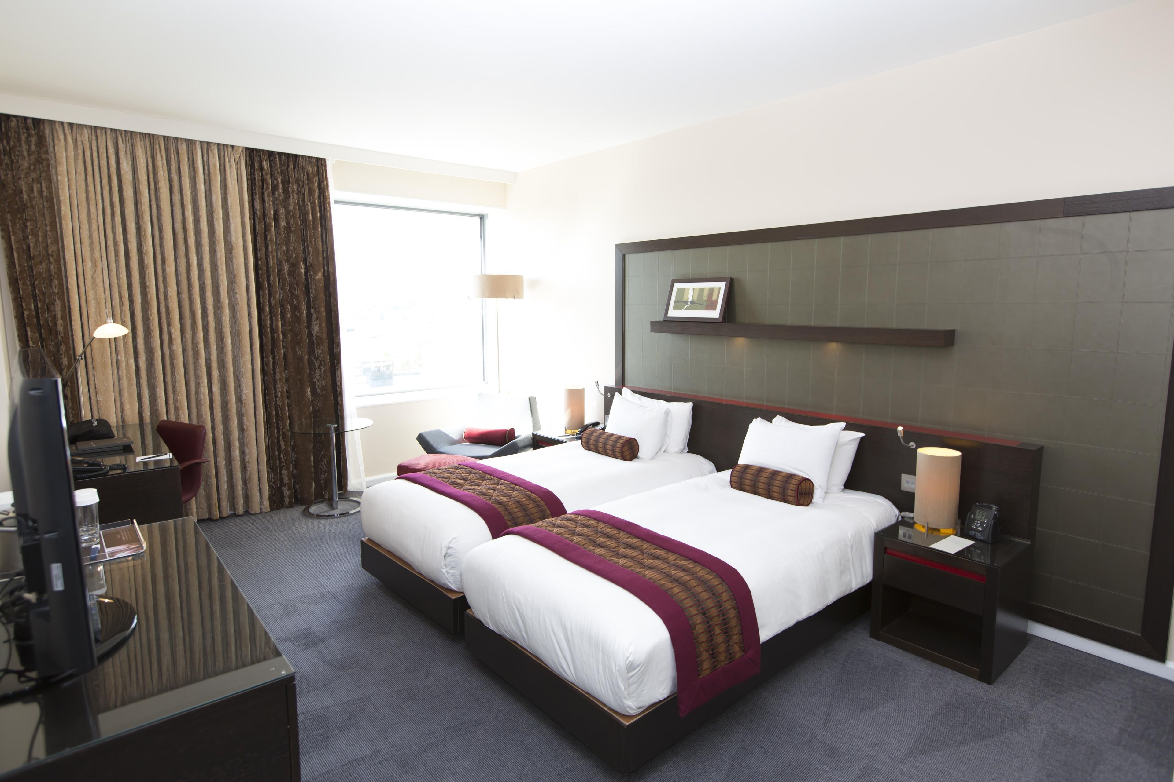 Ein Hotelzimmer mit zwei Betten, einem Schreibtisch, einem Stuhl, einem Fernseher und einem Fenster mit Vorhängen.