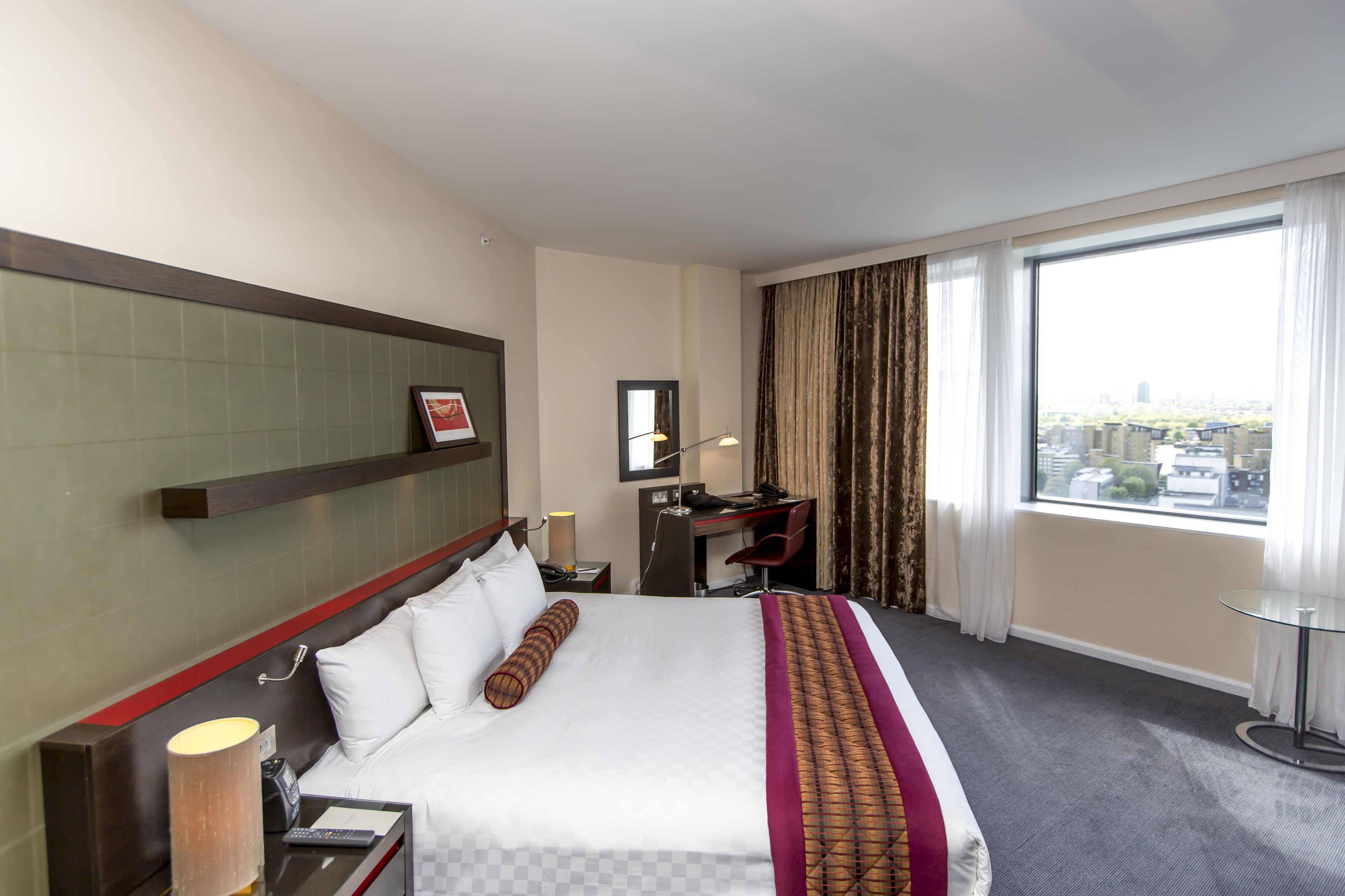 Habitación de hotel con cama, escritorio, silla, lámpara, espejo y ventana con cortinas.