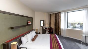 Habitación de hotel con cama, escritorio, silla, lámpara, espejo y ventana con cortinas.
