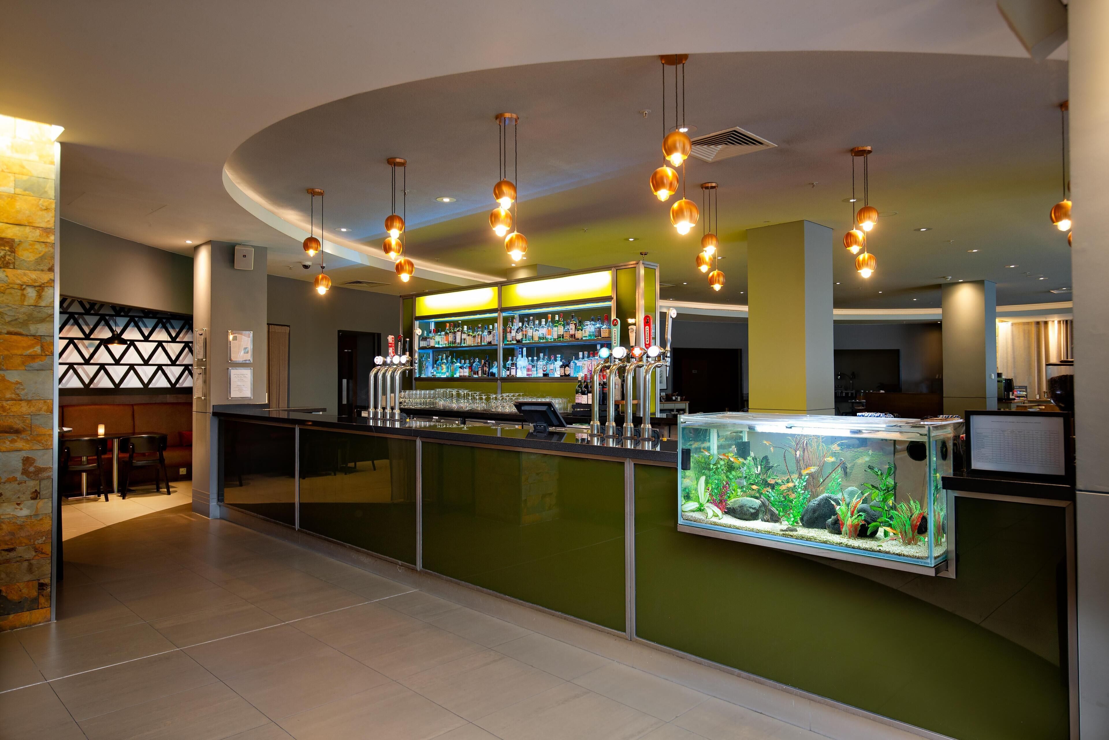 A bar area with a green counter, multiple hanging light fixtures, a fish tank, and a bar stocked with bottles.