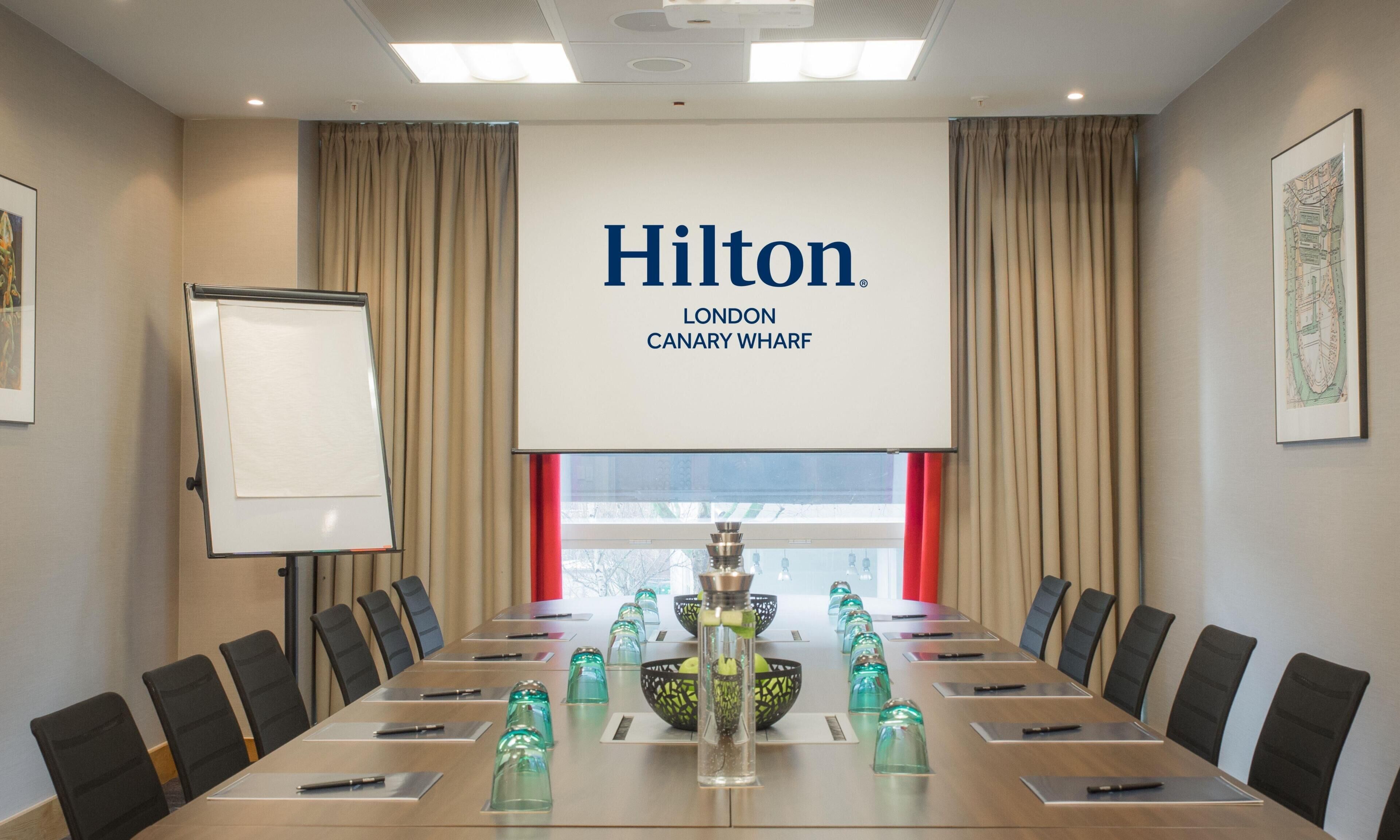 A conference room with a long table, chairs, a projector screen, and a large window with curtains.