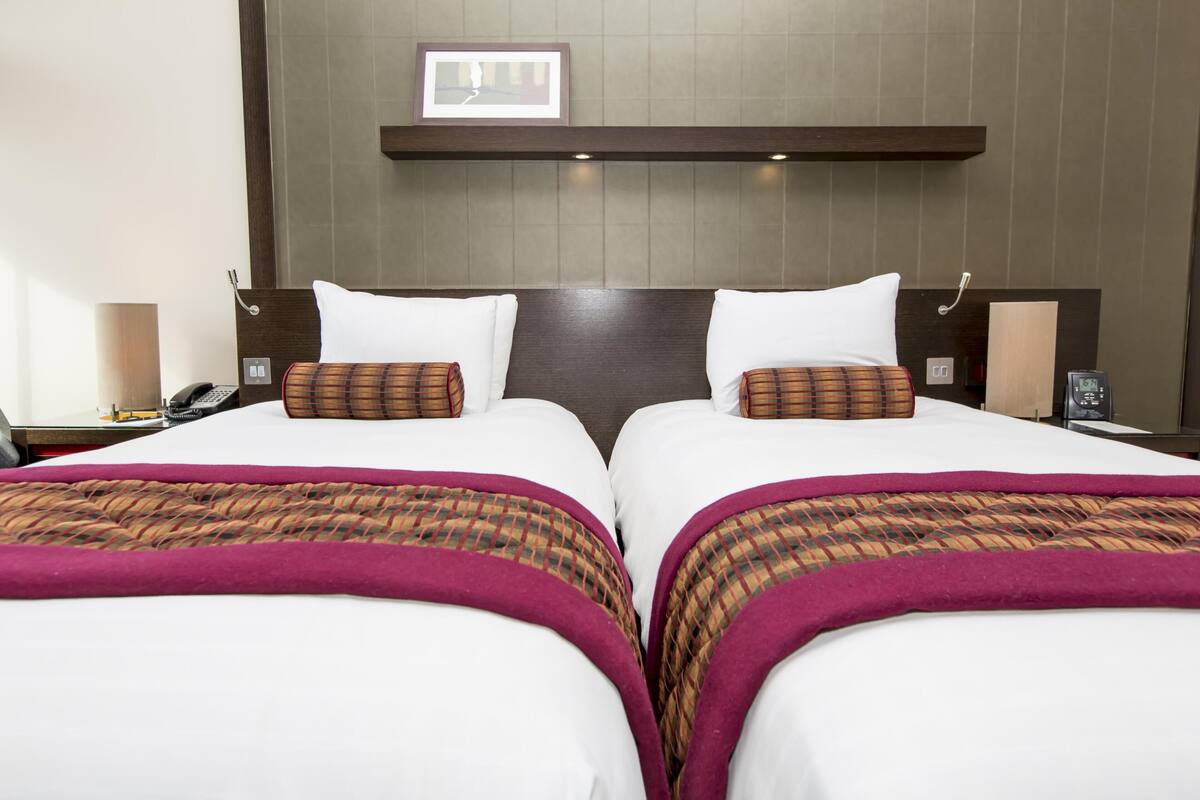 Hotellihuone, jossa on kaksi sänkyä, sängynpääty, yöpöydät, lamppu ja seinällä oleva kehystetty kuva.