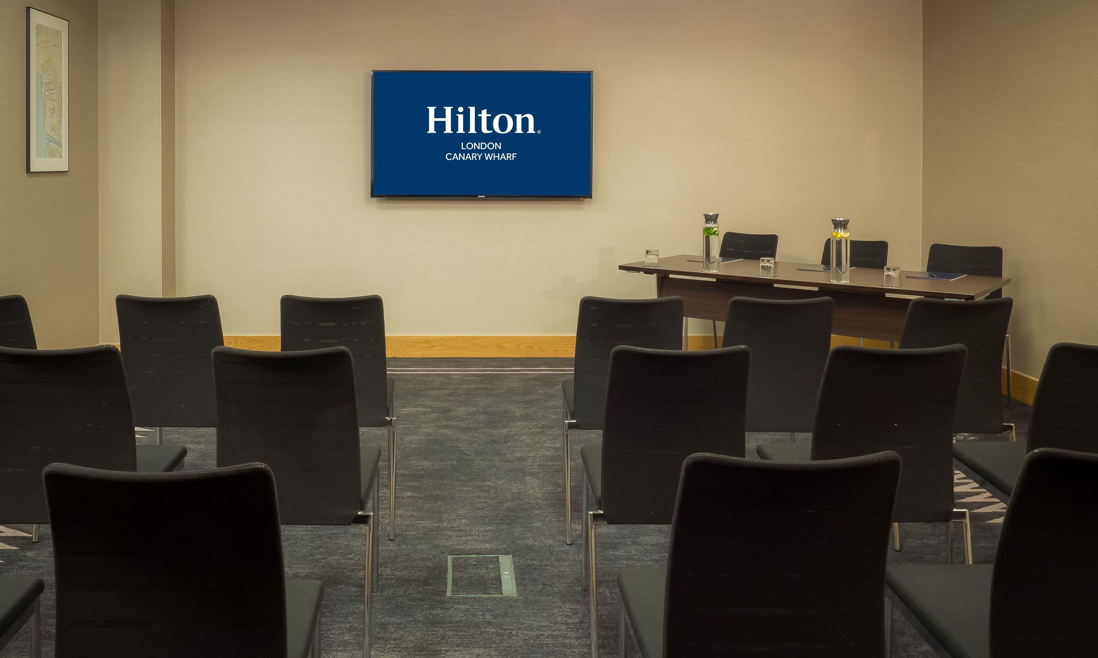 A conference room with a Hilton London Canary Wharf sign, black chairs, a table with a laptop, and a glass of water.