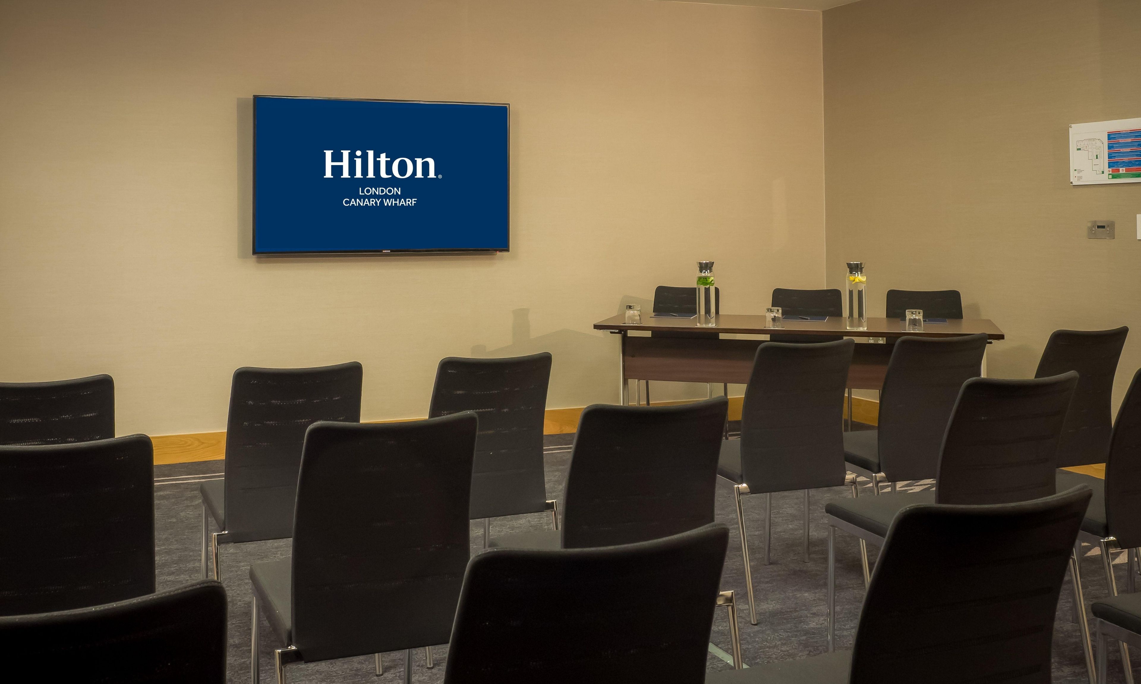 Et mødelokale med et stort skærmbillede, der viser 'Hilton London Canary Wharf', samt et bord med stole og vandflasker.