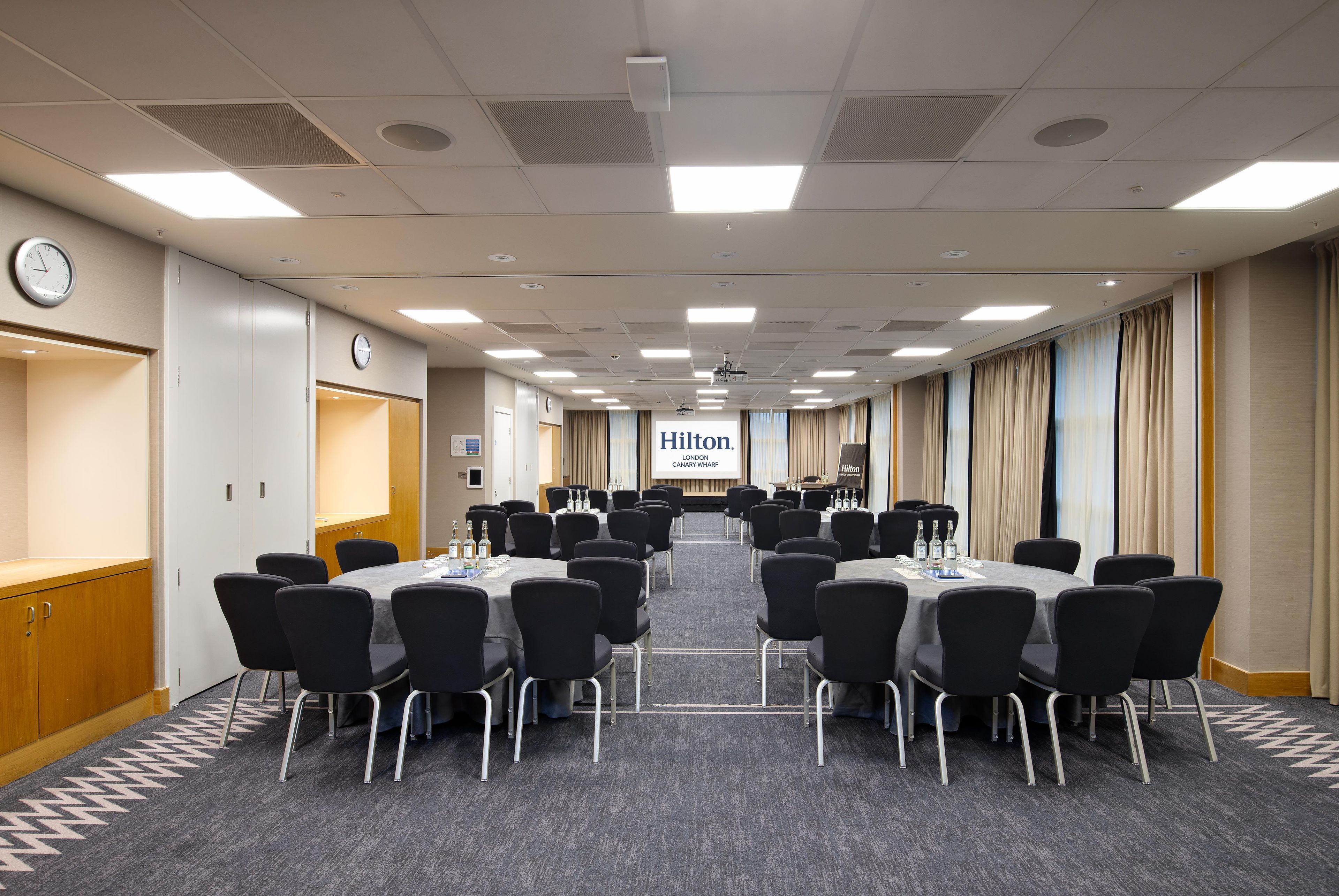 A conference room with multiple tables and chairs, a clock, and a projector screen displaying 'Hilton London Canary Wharf'.
