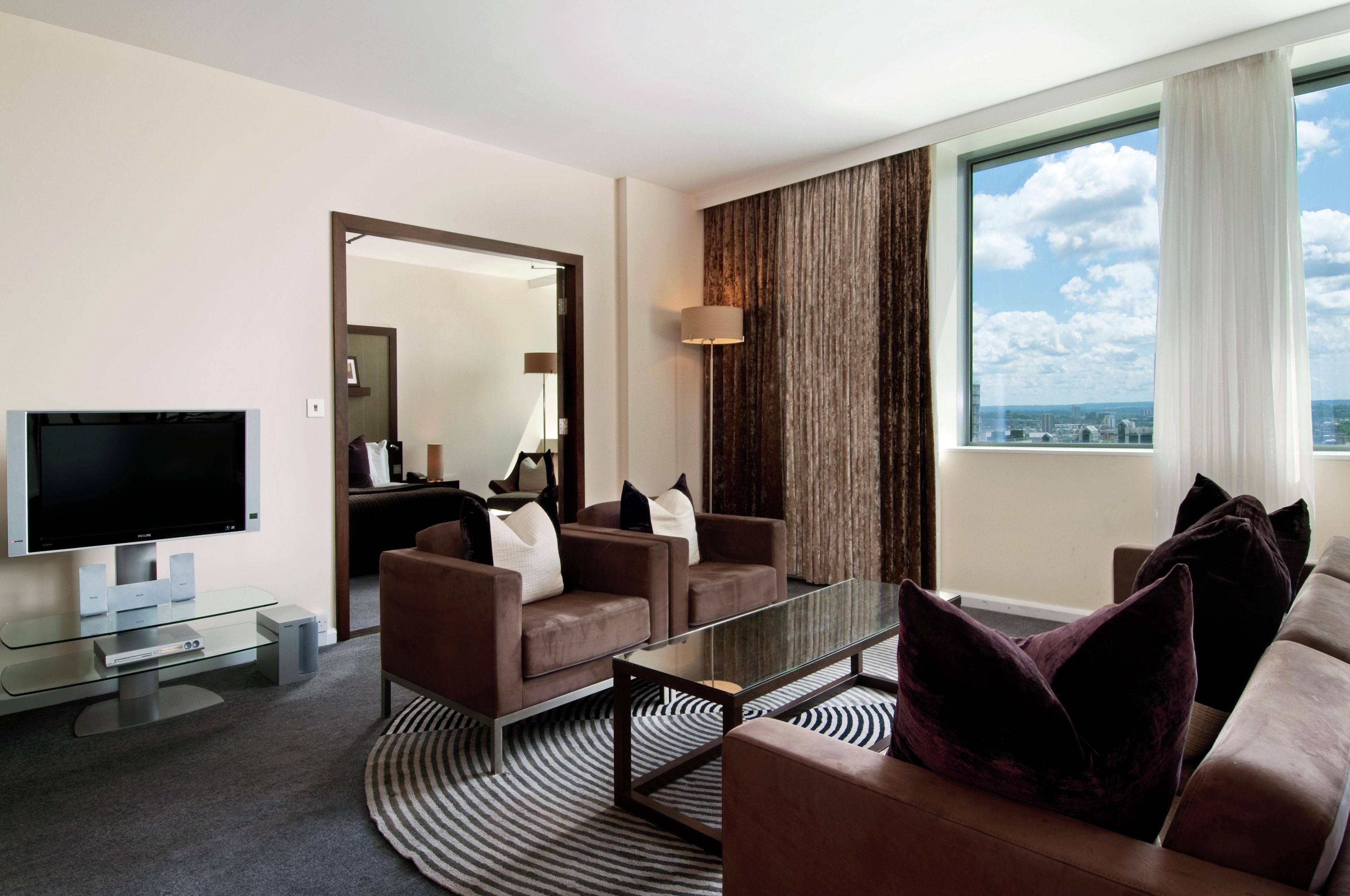 Una habitación de hotel moderna con televisor de pantalla plana, dos sofás, una mesa de centro y un ventanal con vistas a la ciudad.