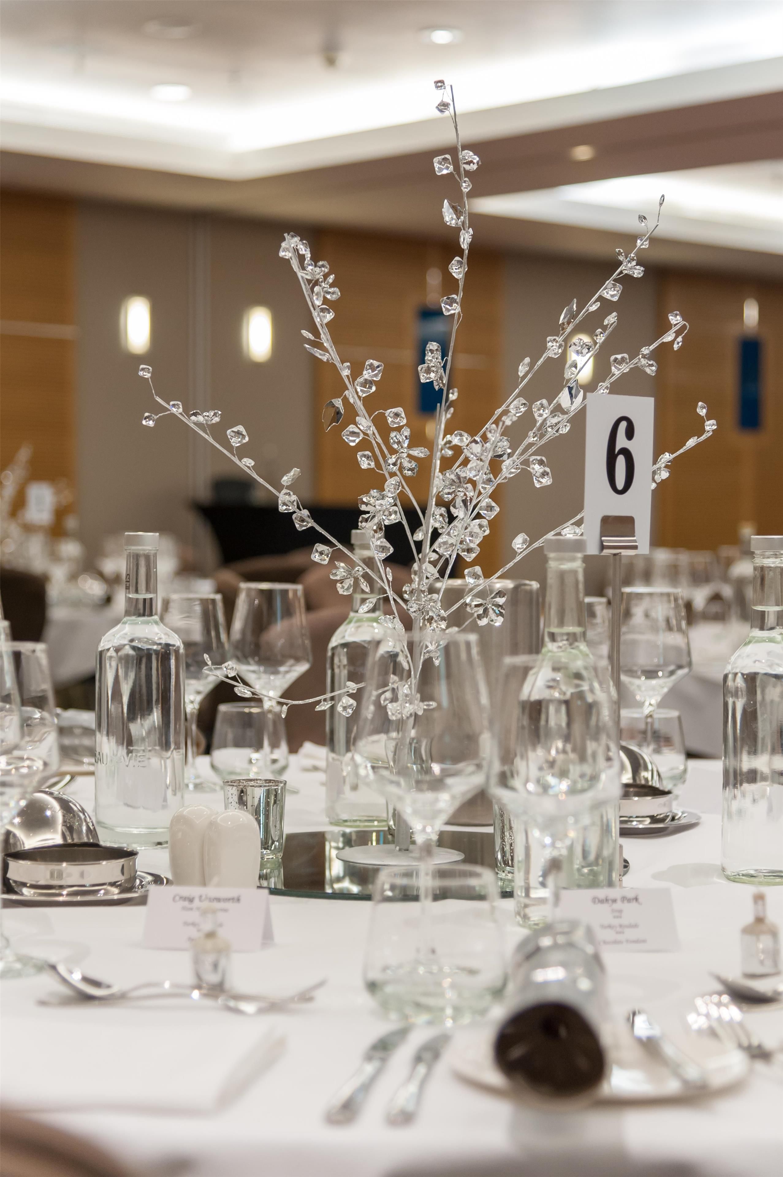 Une table de banquet dressée avec de la verrerie transparente, des marque-places numérotés et une branche de cristal décorative en guise de centre de table.