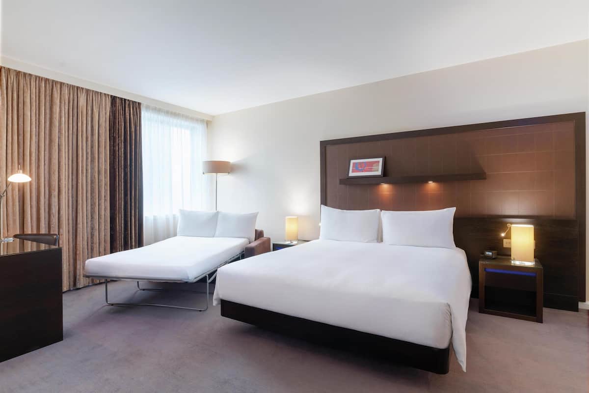Hotellihuoneessa on suuri sänky, pienempi sänky, työpöytä ja yöpöytä, jossa on lamppu.