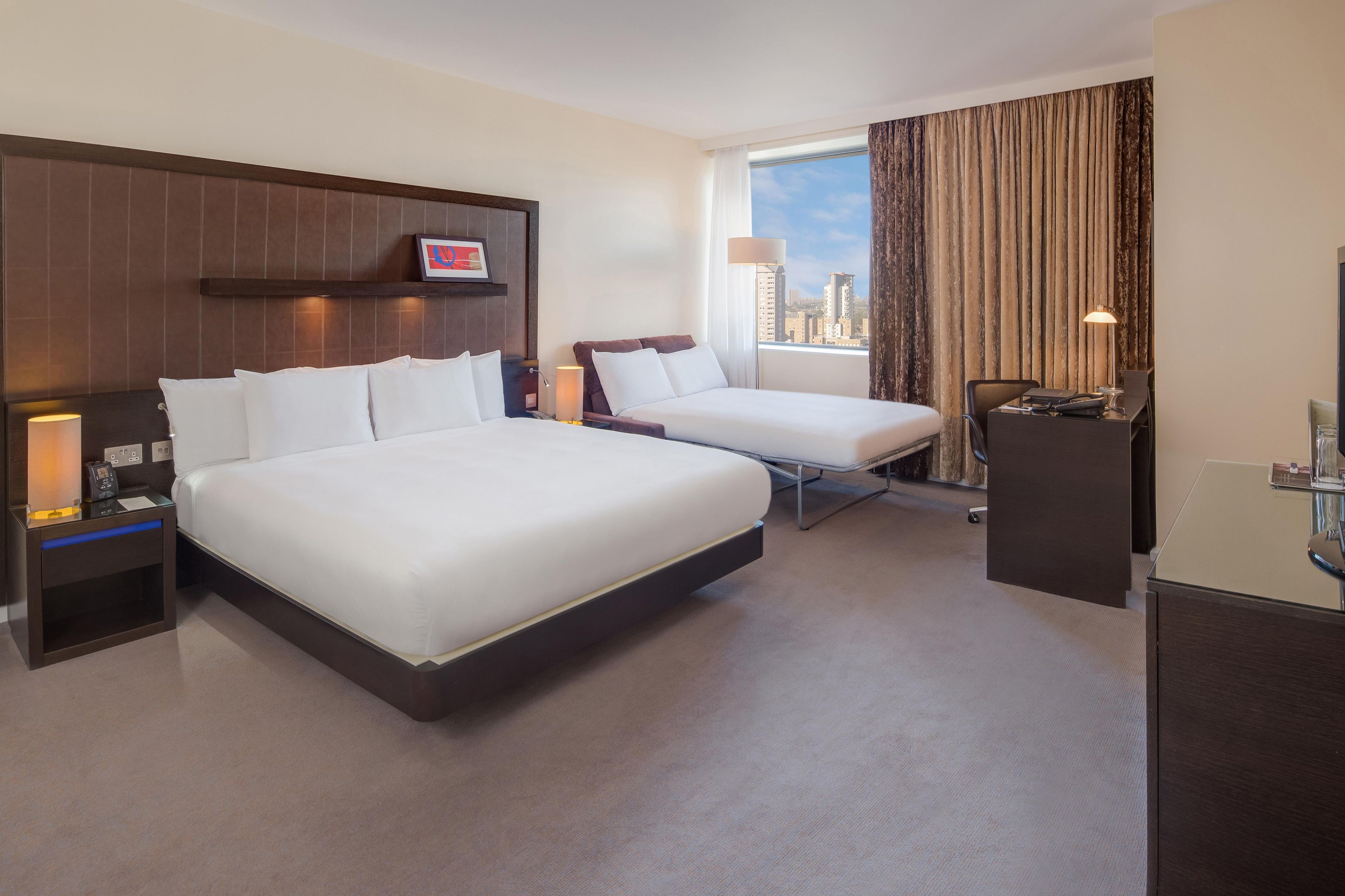 A hotel room with two beds, a desk, a chair, a lamp, and a city view.