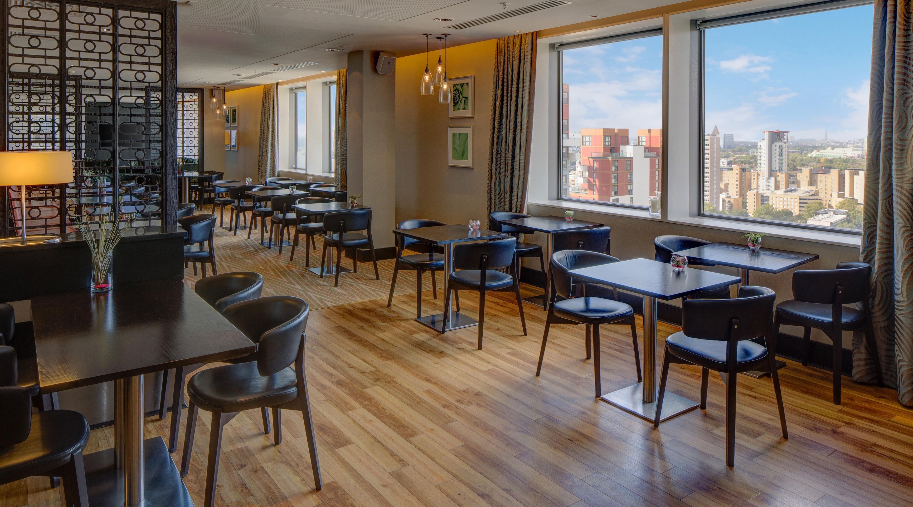 Moderni ravintola, jossa on puulattiat, mustat tuolit ja pöydät. Suuret ikkunat tarjoavat näkymän kaupunkiin.