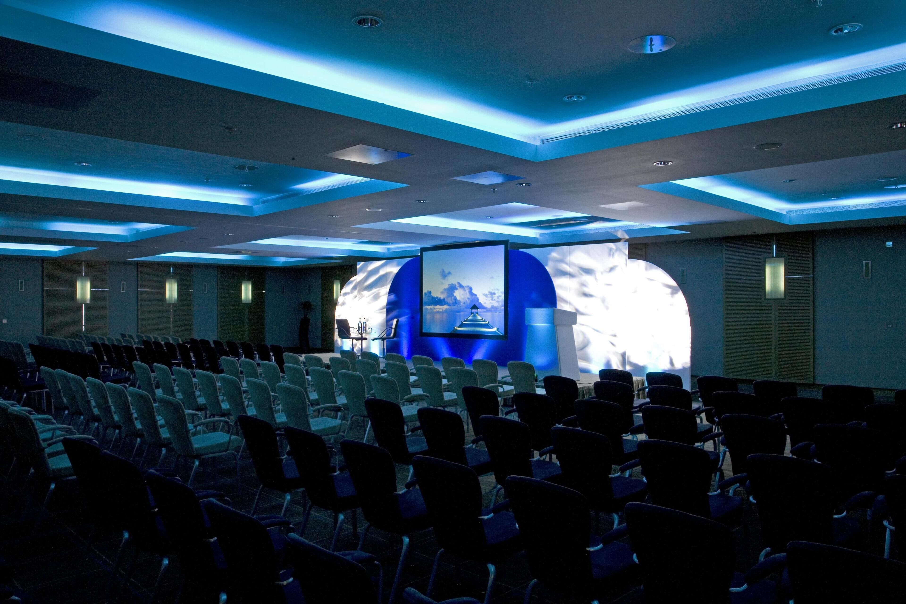 A conference room with rows of chairs, a large screen, and blue ceiling lighting.