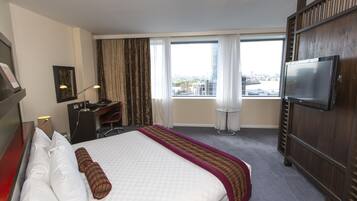 A hotel room with a bed, desk, chair, TV, and large windows with curtains.
