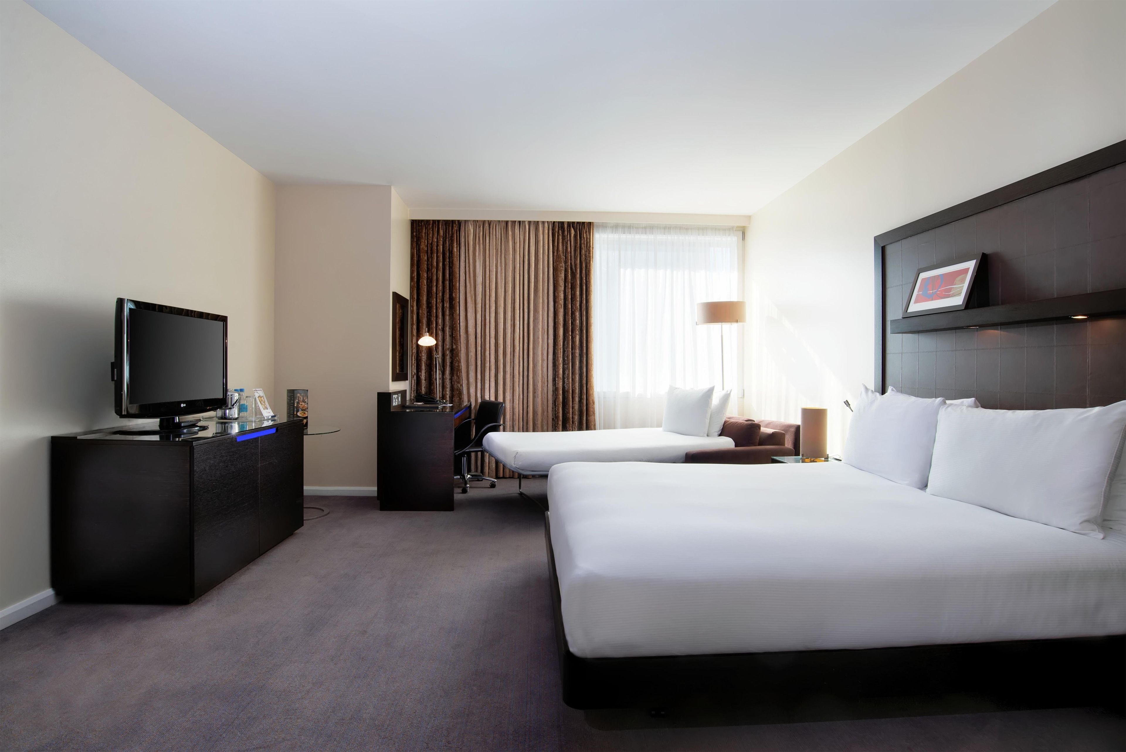 A hotel room with a bed, a desk with a TV, a chair, a lamp, and a window with curtains.
