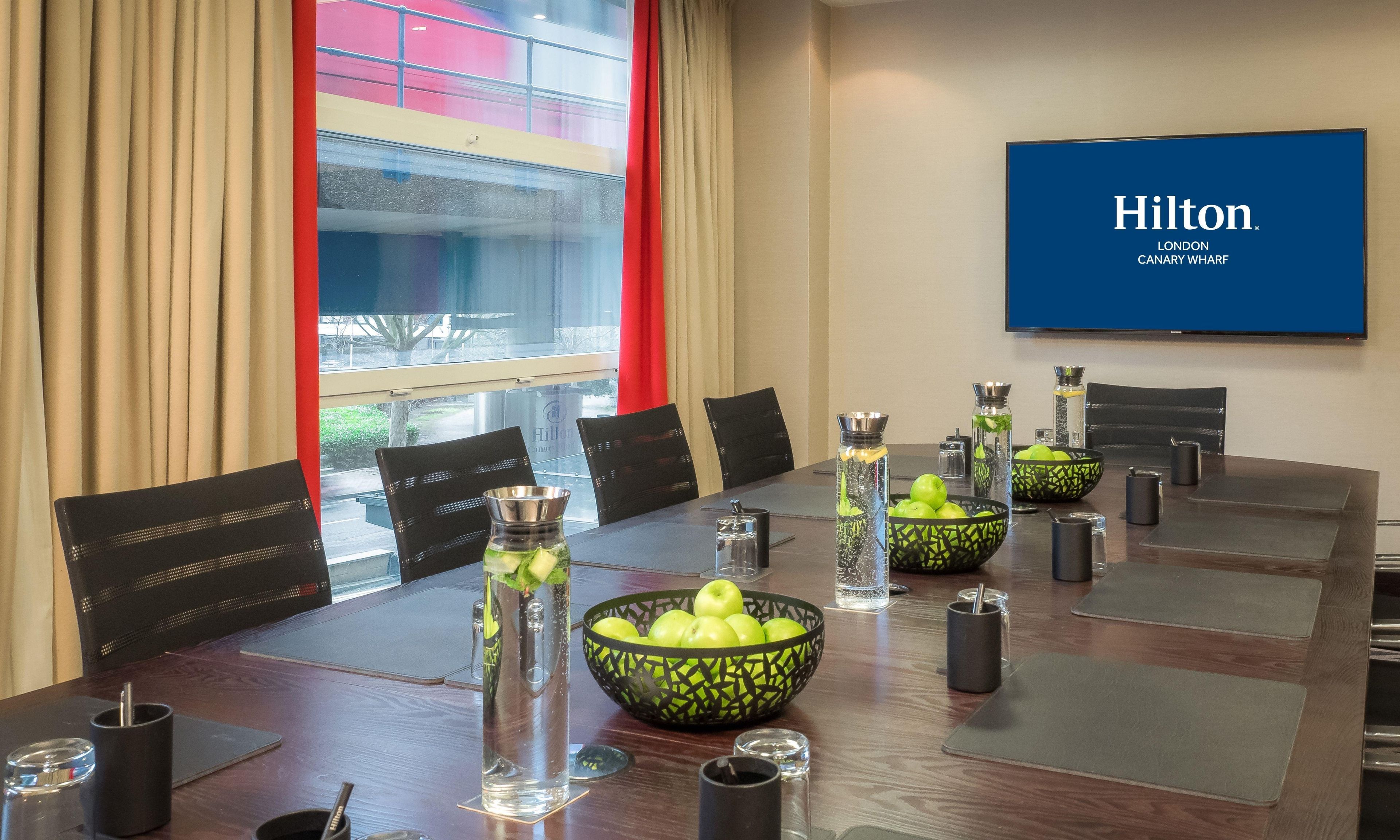 A conference room with a long table, black chairs, a large screen displaying 'Hilton London Canary Wharf', and decorative items including vases and bowls of apples.