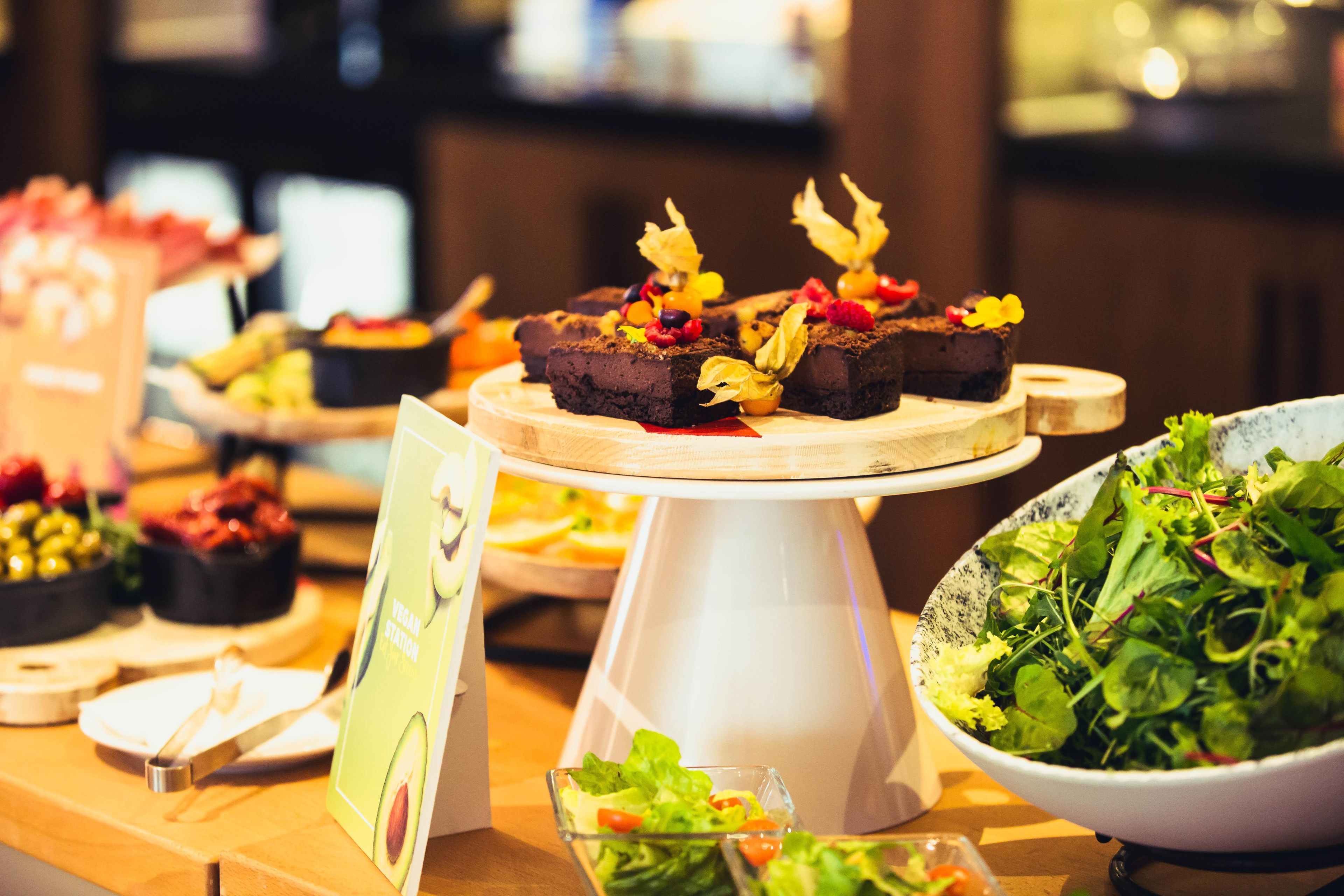 Una mesa de buffet con diversos alimentos, incluyendo un bol de ensalada, un plato de brownies cubiertos de frutas y un letrero que indica opciones veganas.