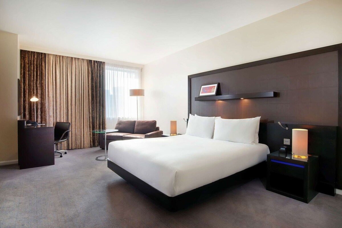 A hotel room with a large bed, a desk with a chair, a sofa, a nightstand with a lamp, and a wall-mounted TV.