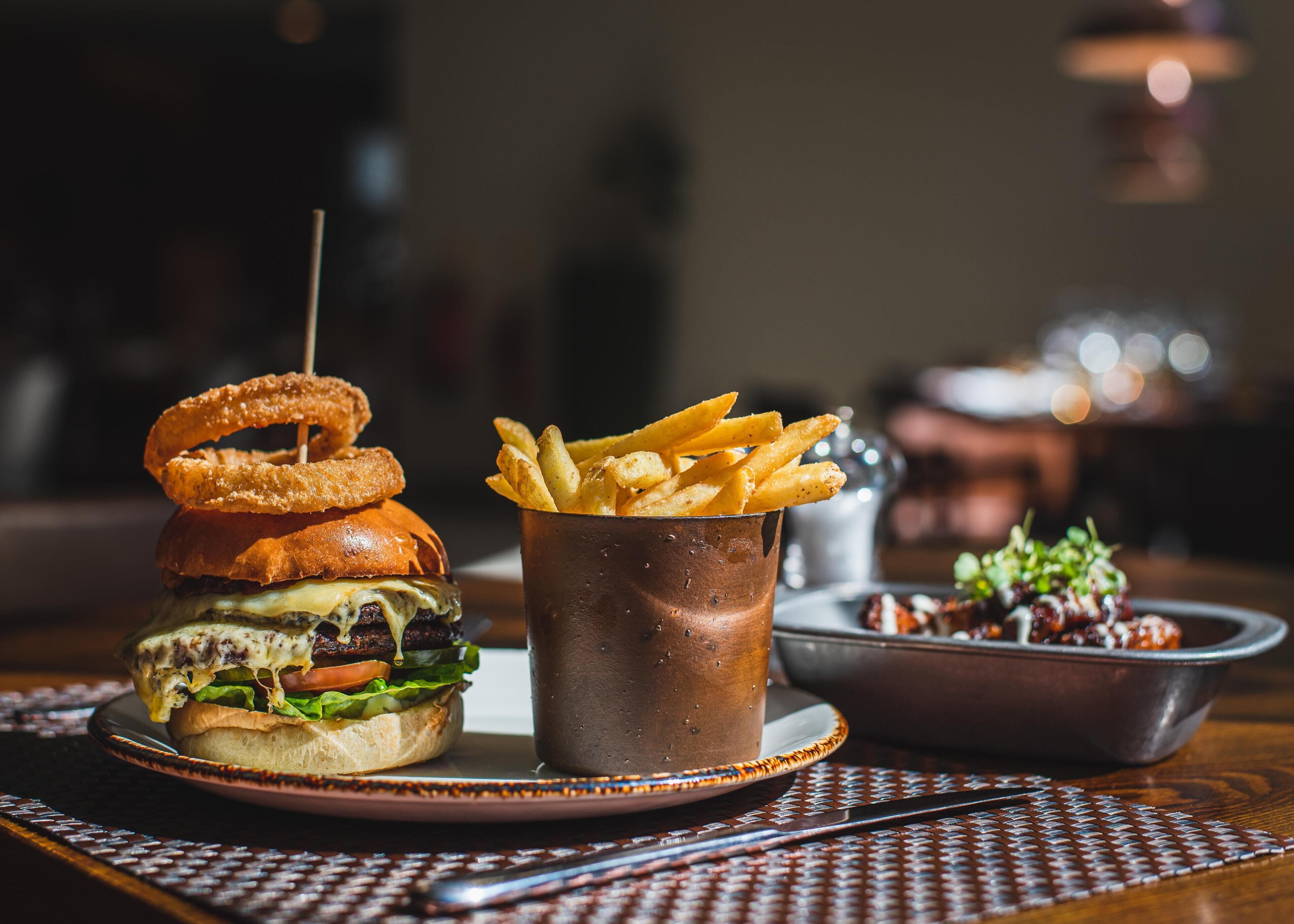 Hamburguesa con aros de cebolla y papas fritas en un plato, con un vaso metálico y un bol de ensalada al fondo.