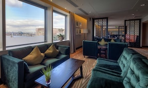 A hotel lobby with a large window offering a city view, a seating area with green upholstered sofas, and a small table with a plant. - Hilton London Canary Wharf (London)