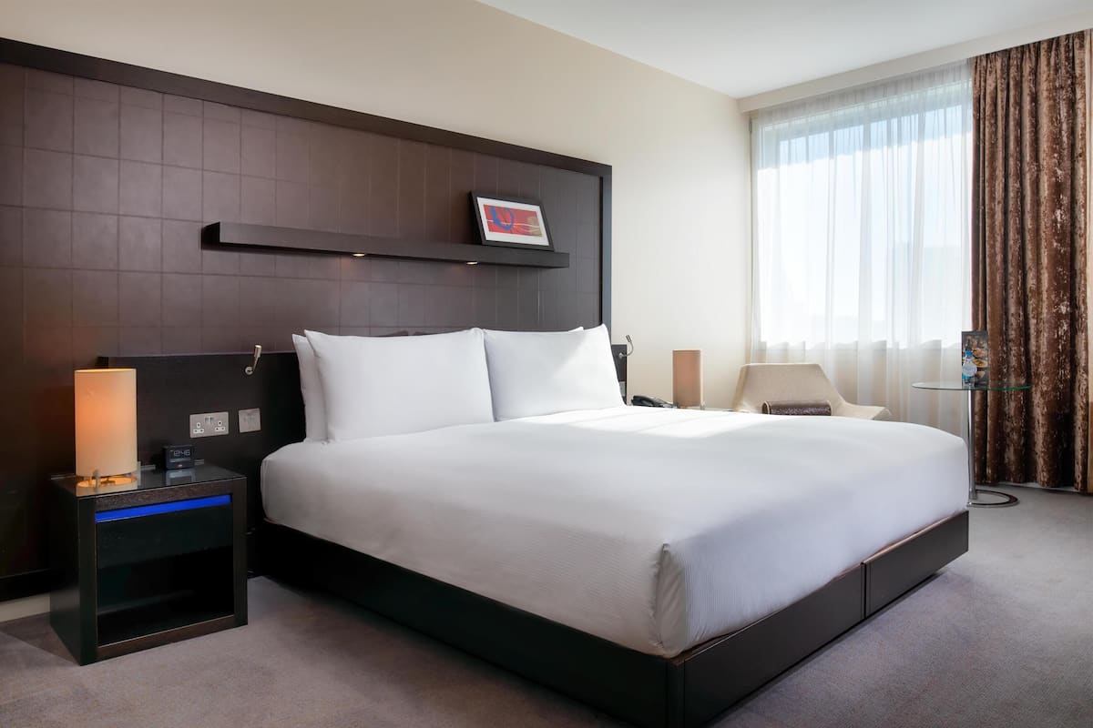 Hotellihuone, jossa on suuri sänky, yöpöytä lampun kanssa, pieni pöytä lampun kanssa ja ikkuna, jossa on verhot.