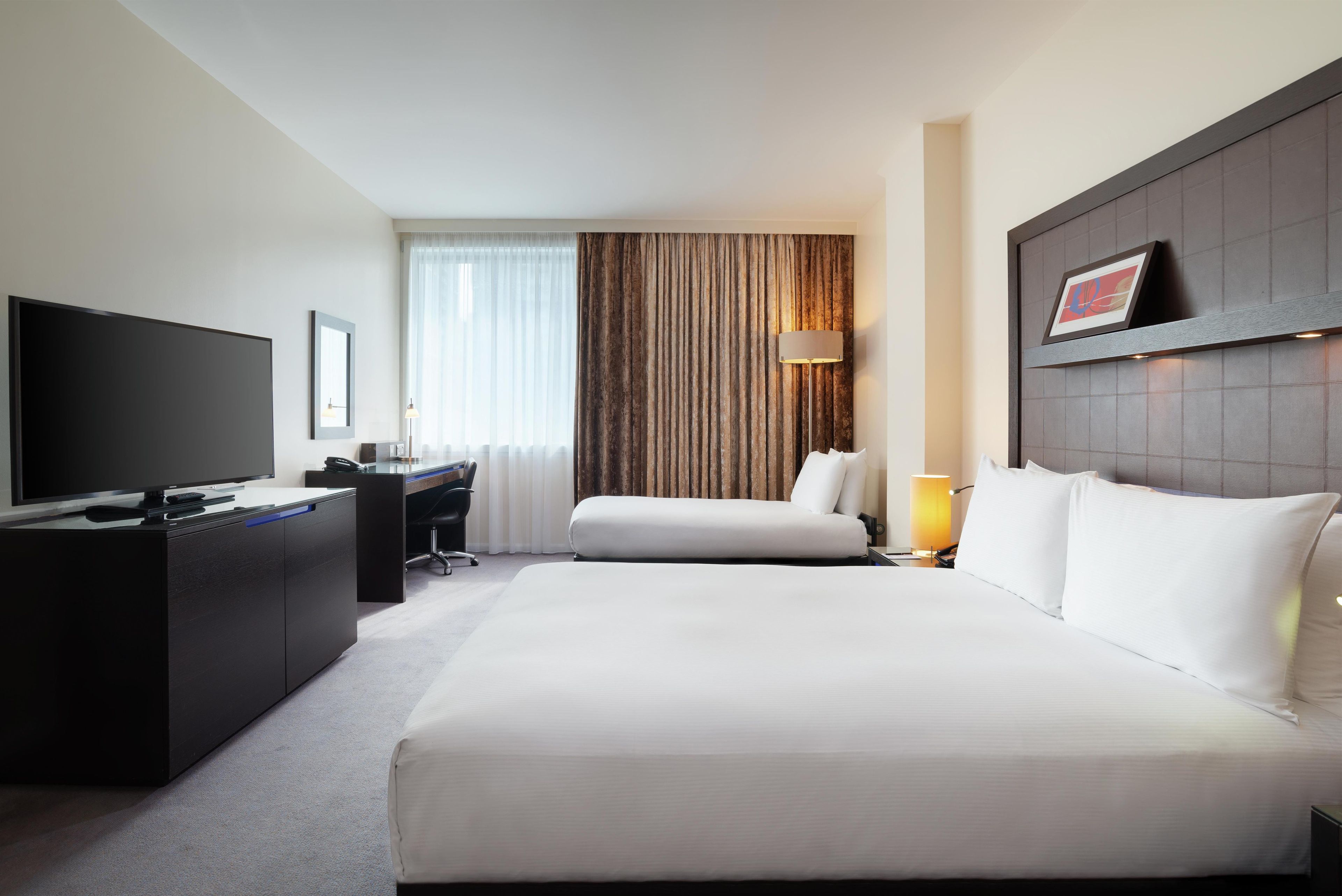 Quarto de hotel com uma cama grande, uma mesa de trabalho com televisão de tela plana, uma cadeira, um abajur e uma janela com cortinas.
