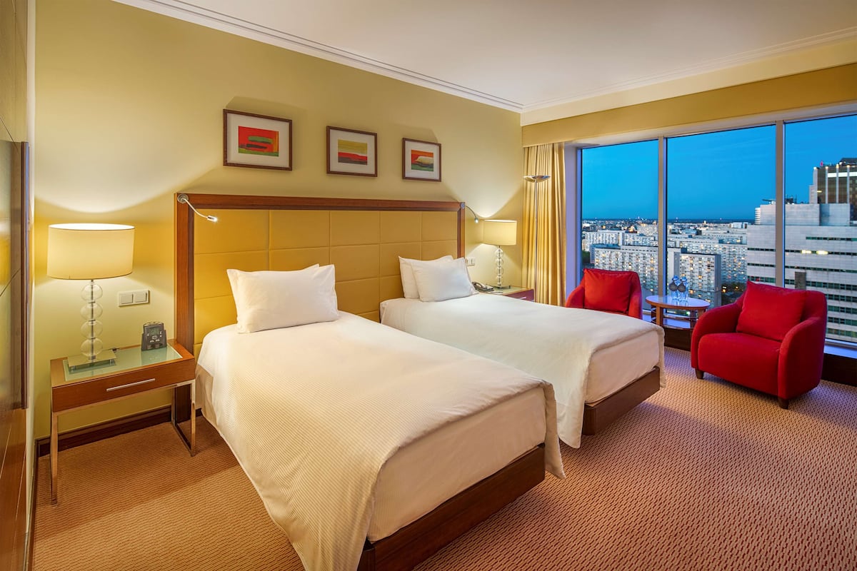 A hotel room with two beds, a red chair, and a large window offering a city view.
