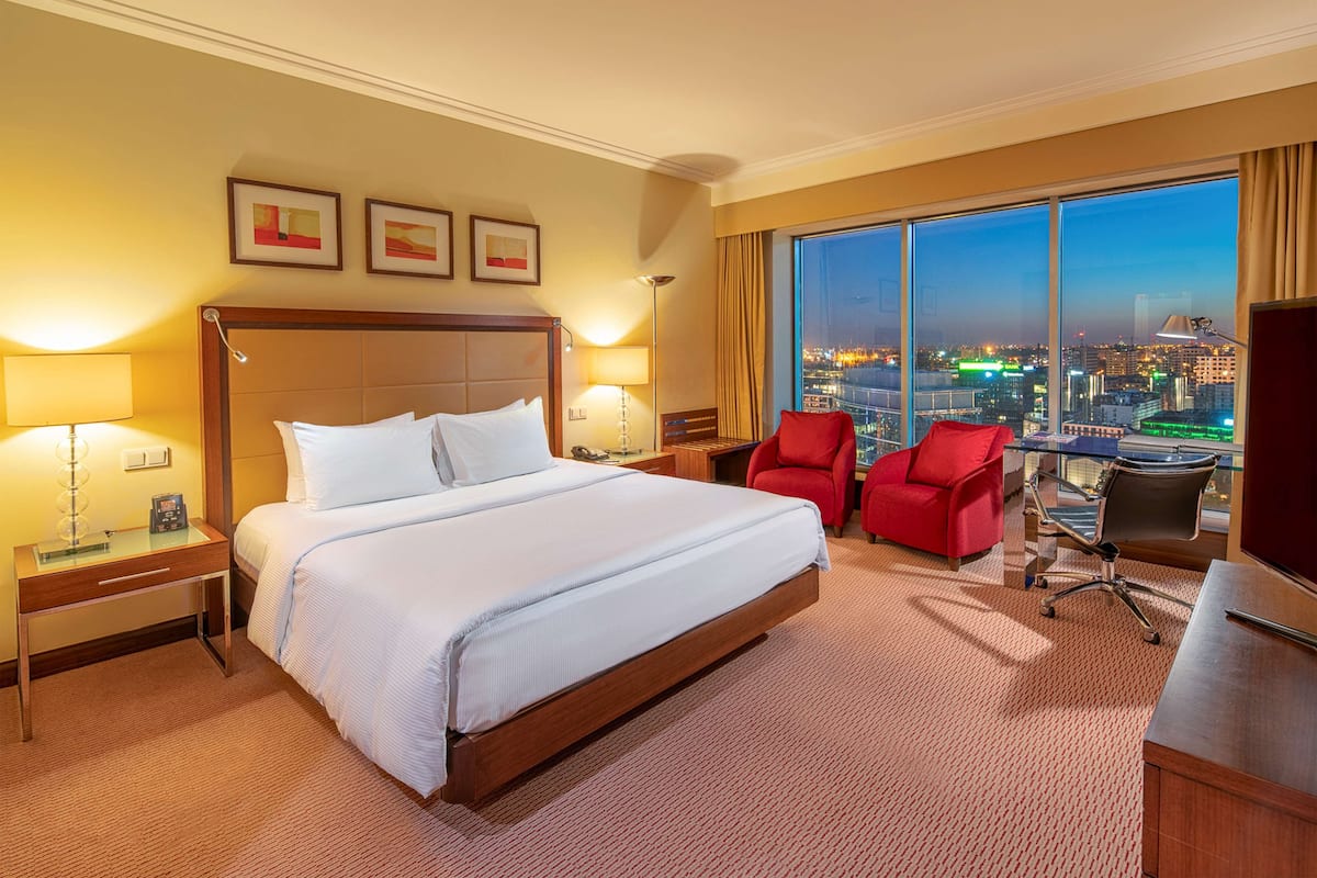 A hotel room with a large bed, two bedside tables with lamps, a desk with a chair, and a large window offering a city view.