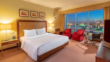 Habitación de hotel con una cama grande, dos mesitas de noche con lámparas, un escritorio con silla y un ventanal con vistas a la ciudad.