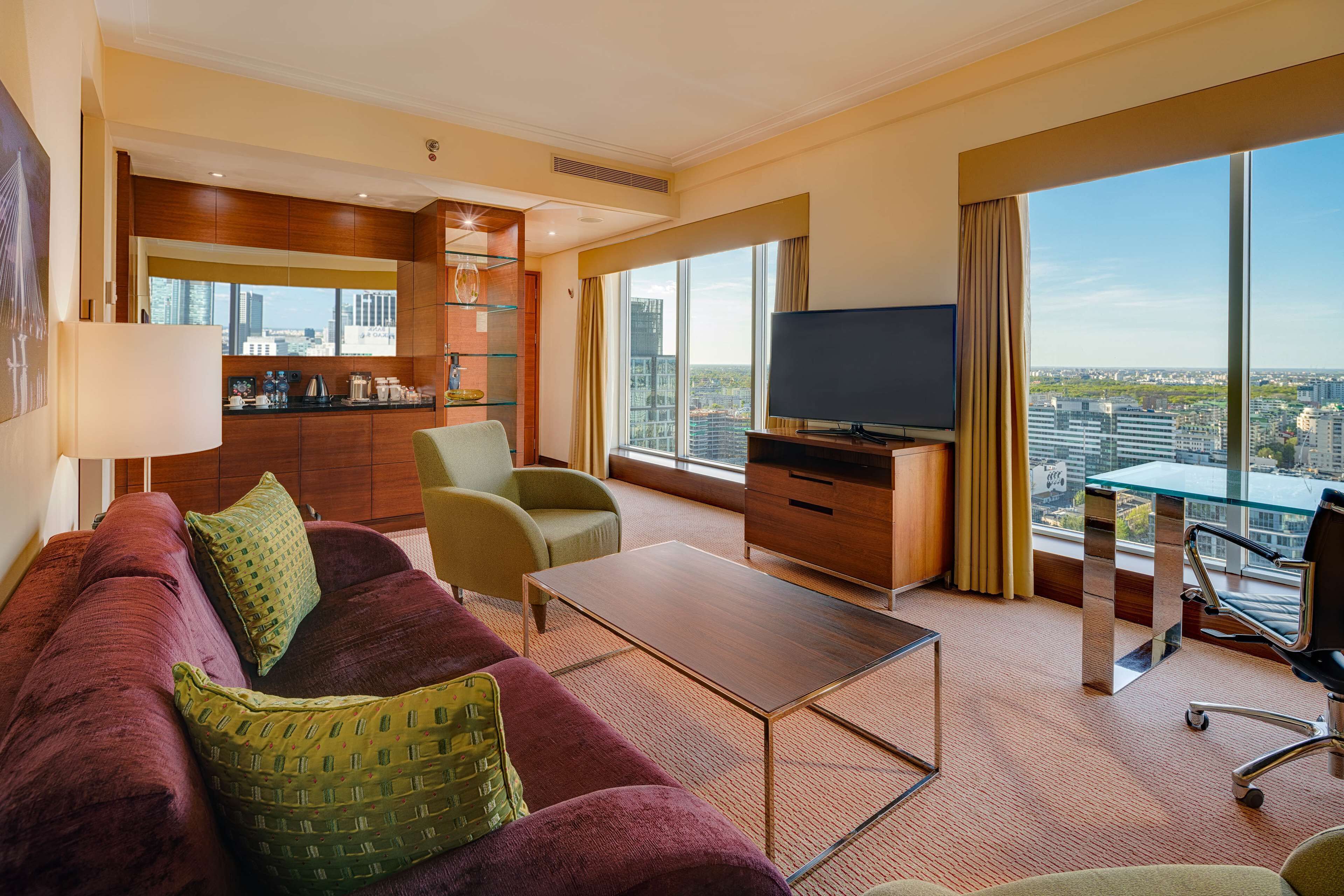 A modern hotel room with a large window offering a city view, a plush red sofa with green cushions, a wooden desk, and a flat-screen TV.