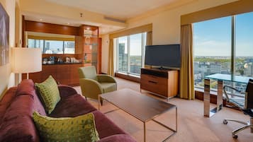 Habitación de hotel moderna con un gran ventanal que ofrece vistas a la ciudad, un sofá rojo cómodo con cojines verdes, un escritorio de madera y un televisor de pantalla plana.