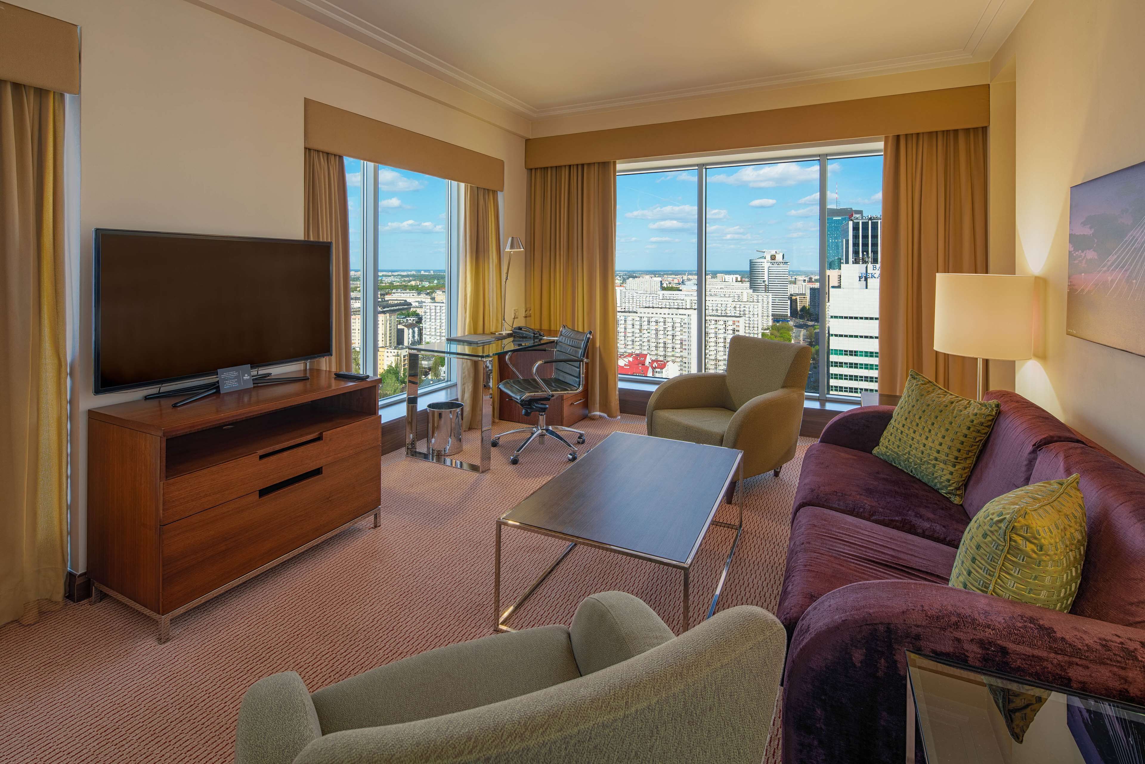 Habitación de hotel con un televisor grande, un sofá, una mesita de centro, un escritorio con una silla y ventanas amplias que ofrecen vista a la ciudad.