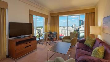Habitación de hotel con un televisor grande, un sofá, una mesita de centro, un escritorio con una silla y ventanas amplias que ofrecen vista a la ciudad.